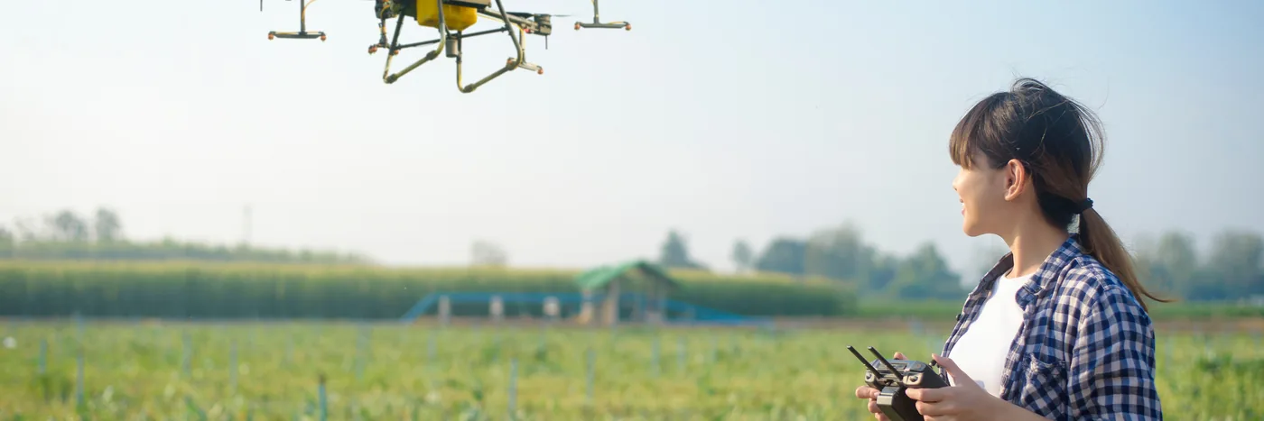 An operator holding a twin-stick controller in a field while a spray drone hovers behind her.