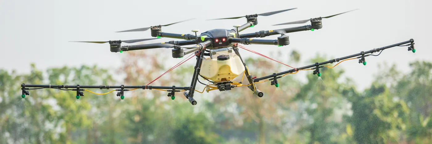 A large multirotor spray drone with a yellow tank hovering low over a green field, booms extended.