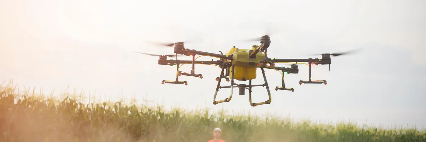 A spray drone seen head-on in flight above a field, tank full and spray booms spread wide.